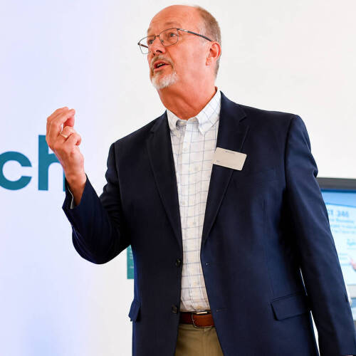 Paul Hillman stands with right arm raised during a presentation at the front of a room